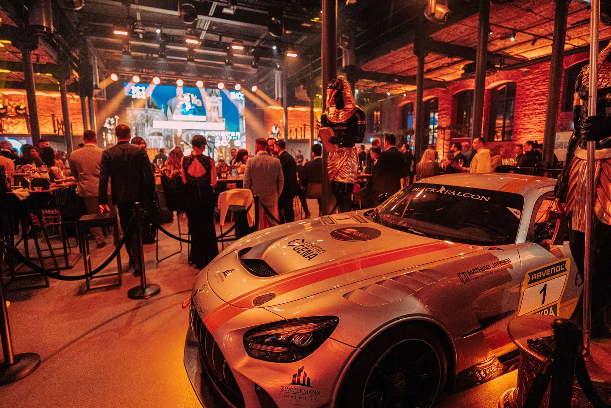 Sportwagen im Vordergrund bei einer Galaveranstaltung. Im Hintergrund sind Gäste vor einer Bühne zu sehen