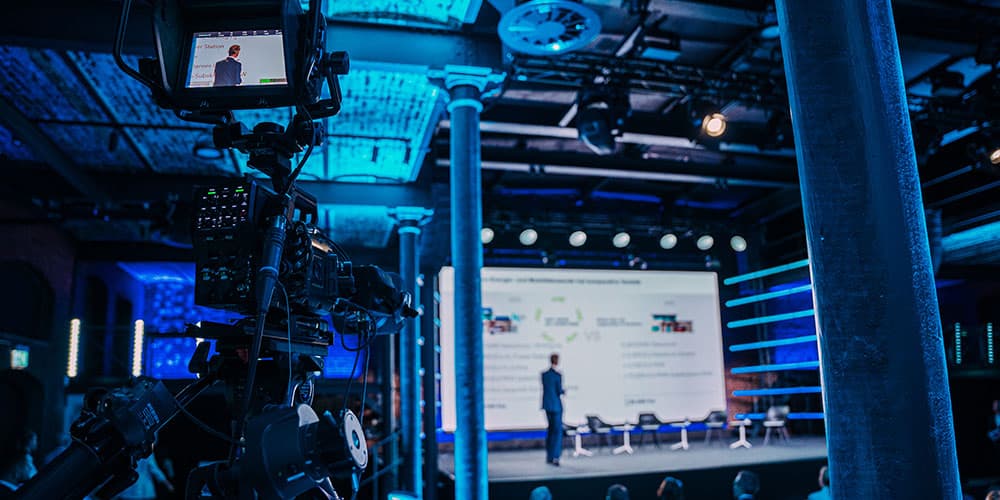 Eine Kamera im Vordergrund bei einer Konferenz. Im Hintergrund ist eine Bühne zu sehen.